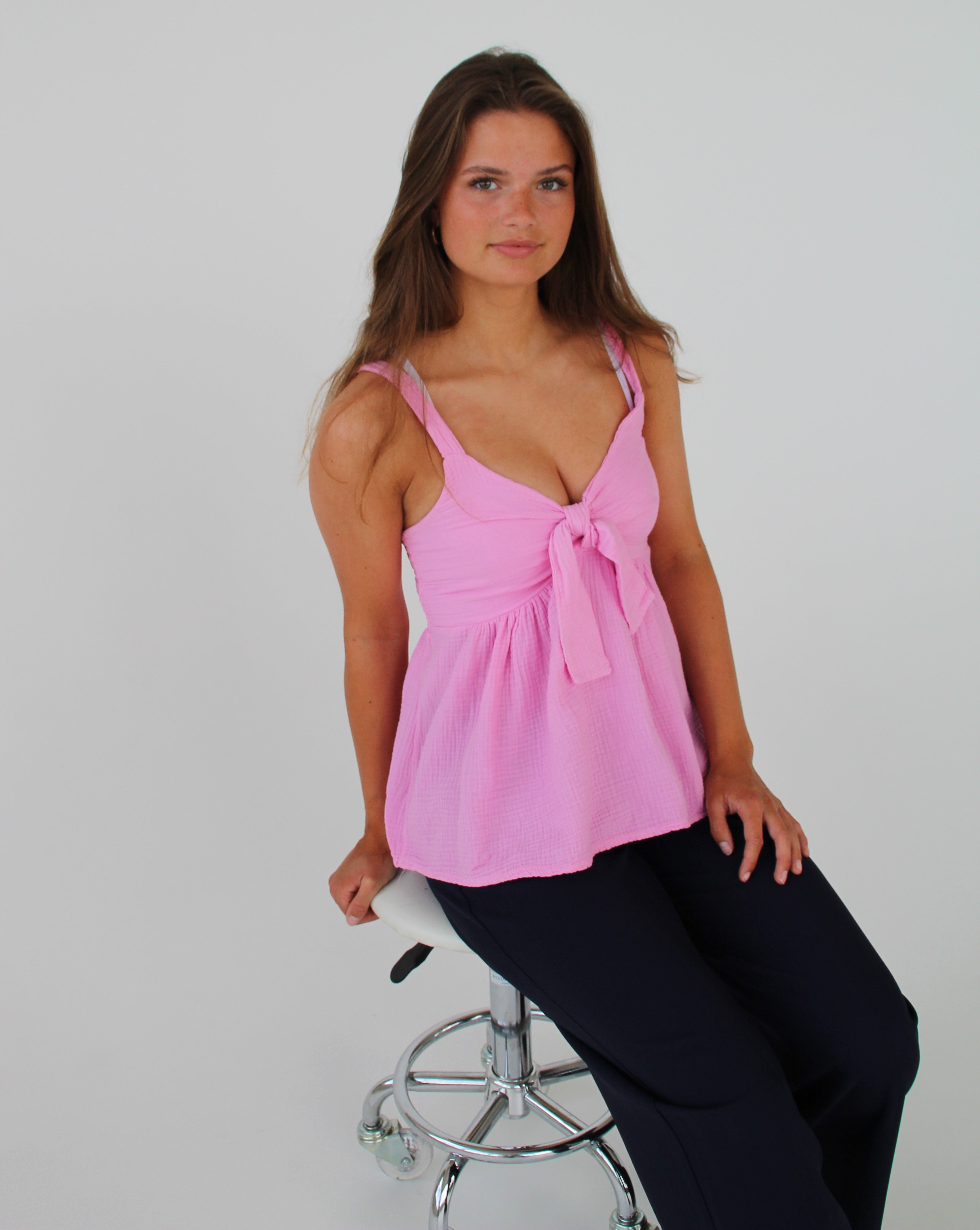 En kvinde sidder på en hvid, polstret taburet iført en lyserød top og mørke bukser. Baggrunden er ensfarvet hvid, hvilket fremhæver hendes positur.