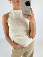 En person iført en cremefarvet top tager et selfiespejlbillede med en mobiltelefon. De har lyse bukser og guldsmykker i et neutralt rum.