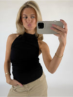 Kvinde tager en selfie med en smartphone, iført sort top og beige bukser. Hun poserer med et ansigtsudtryk og står i et lyst rum.