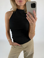 En person fotograferer sig selv med en smartphone iført en sort ærmeløs top og beige bukser. Personen står i et minimalistisk rum og bærer guldsmykker.