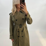 Person iført grøn trenchcoat tager et spejlbillede med en smartphone. Hun står i et minimalistisk rum med lyse vægge.