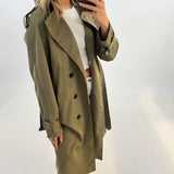 En person iført en grøn trenchcoat tager en selfie i et spejl. Personen bærer også en hvid trøje og bukser i en lys, minimalistisk baggrund.