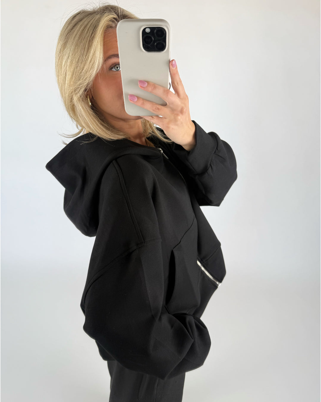 En person iført en sort hættetrøje holder en mobiltelefon op foran ansigtet og tager en selfie i et lyst rum.