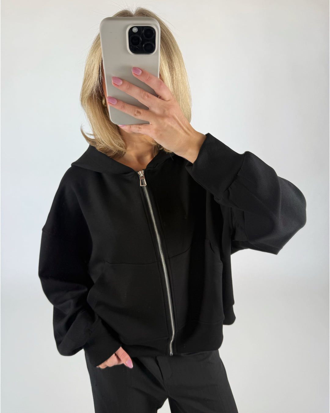 Person iført sort hættetrøje tager et selfie med en smartphone foran ansigtet, mens de står i et rum med enkel, lys baggrund.