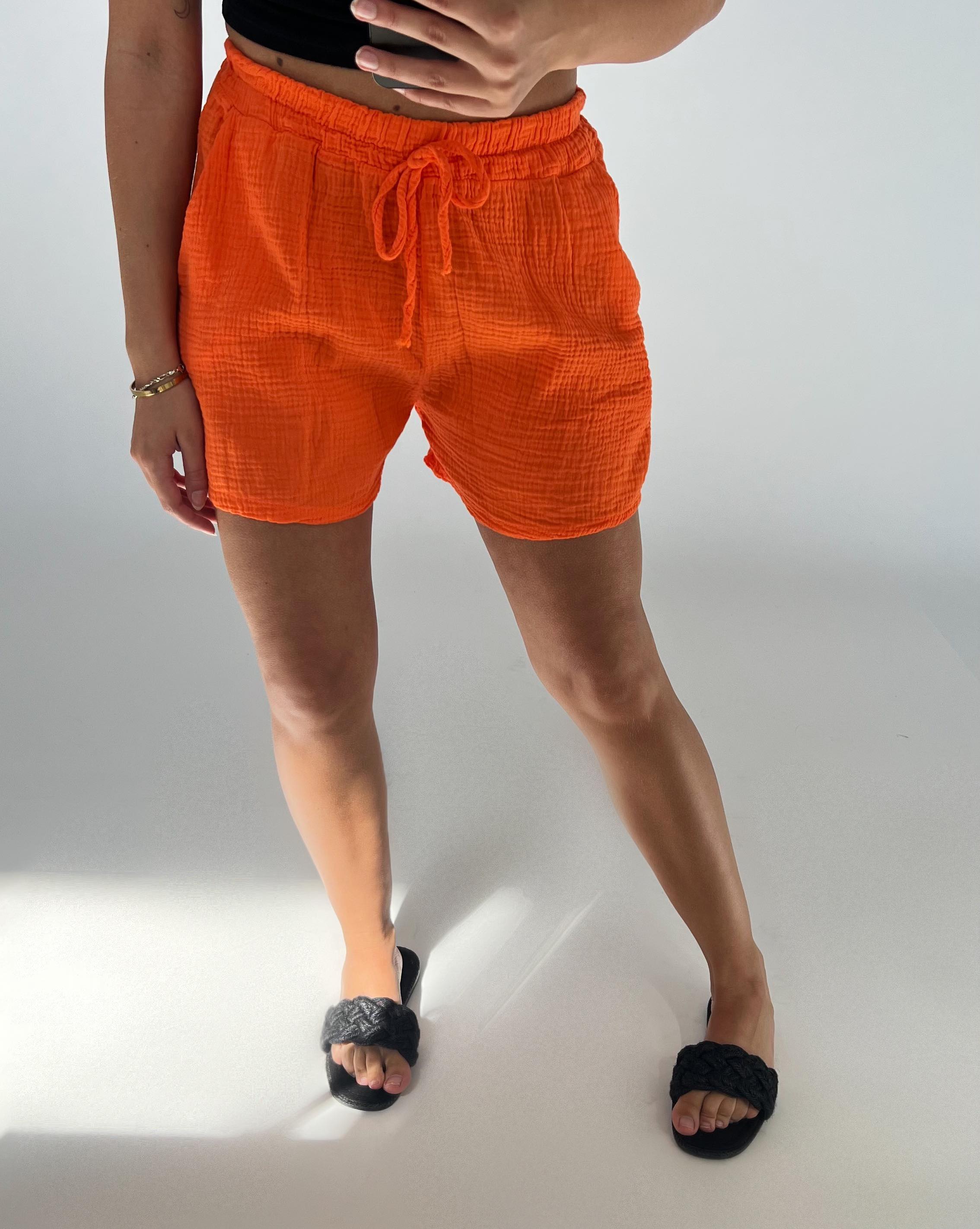En person bærer orange shorts og sorte sandaler i en minimalistisk lys indendørs omgivelser.
