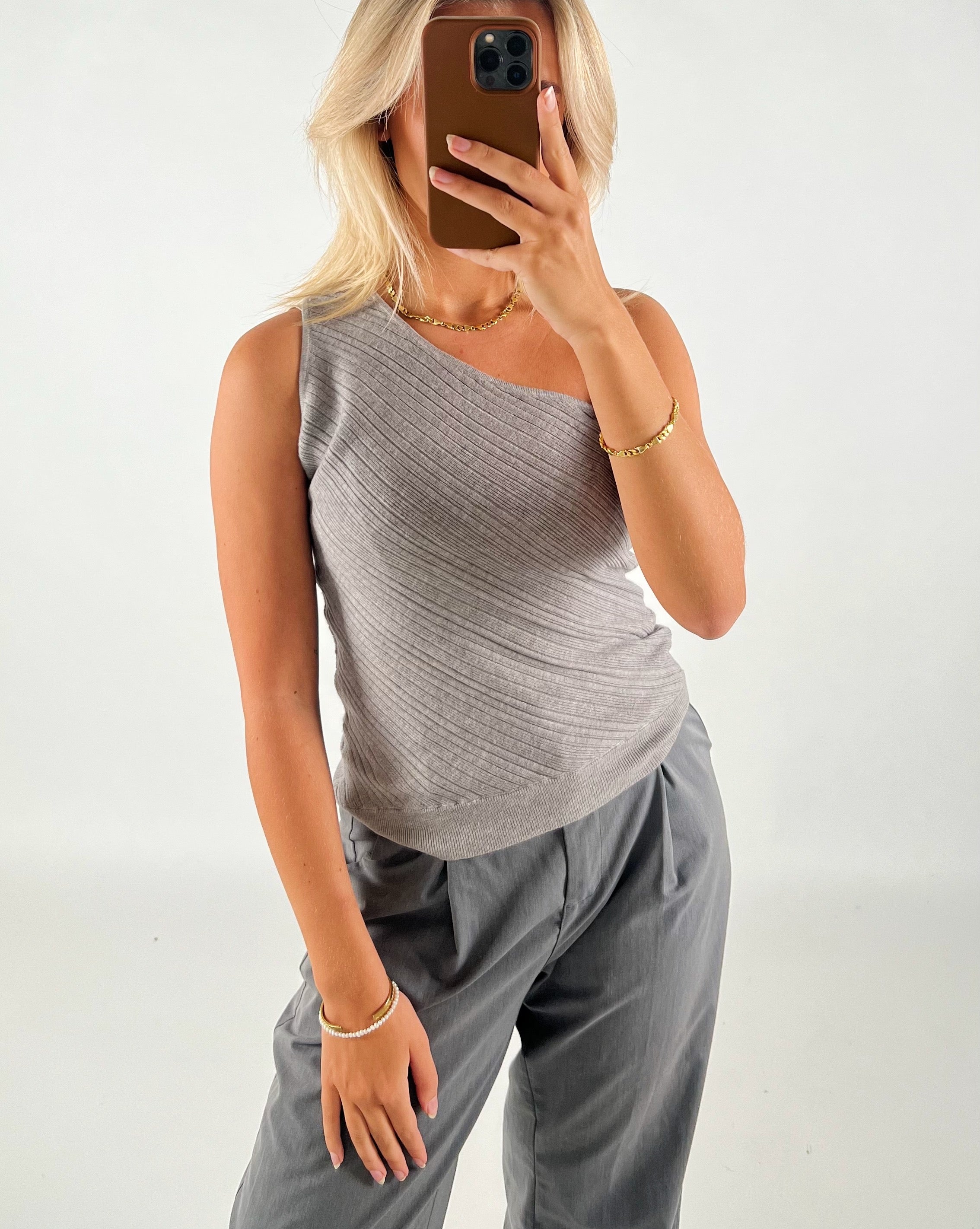 En person iført gråt asymmetrisk bluse og bukser tager en selfie med en smartphone i et minimalistisk rum. Personen har blondt hår og bærer guldsmykker.