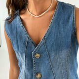 En person iført en ærmeløs denimvest med tre knapper og en perlekæde rundt om halsen står mod en lys baggrund.
