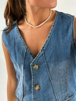 En person iført en ærmeløs denimvest med tre knapper og en perlekæde rundt om halsen står mod en lys baggrund.