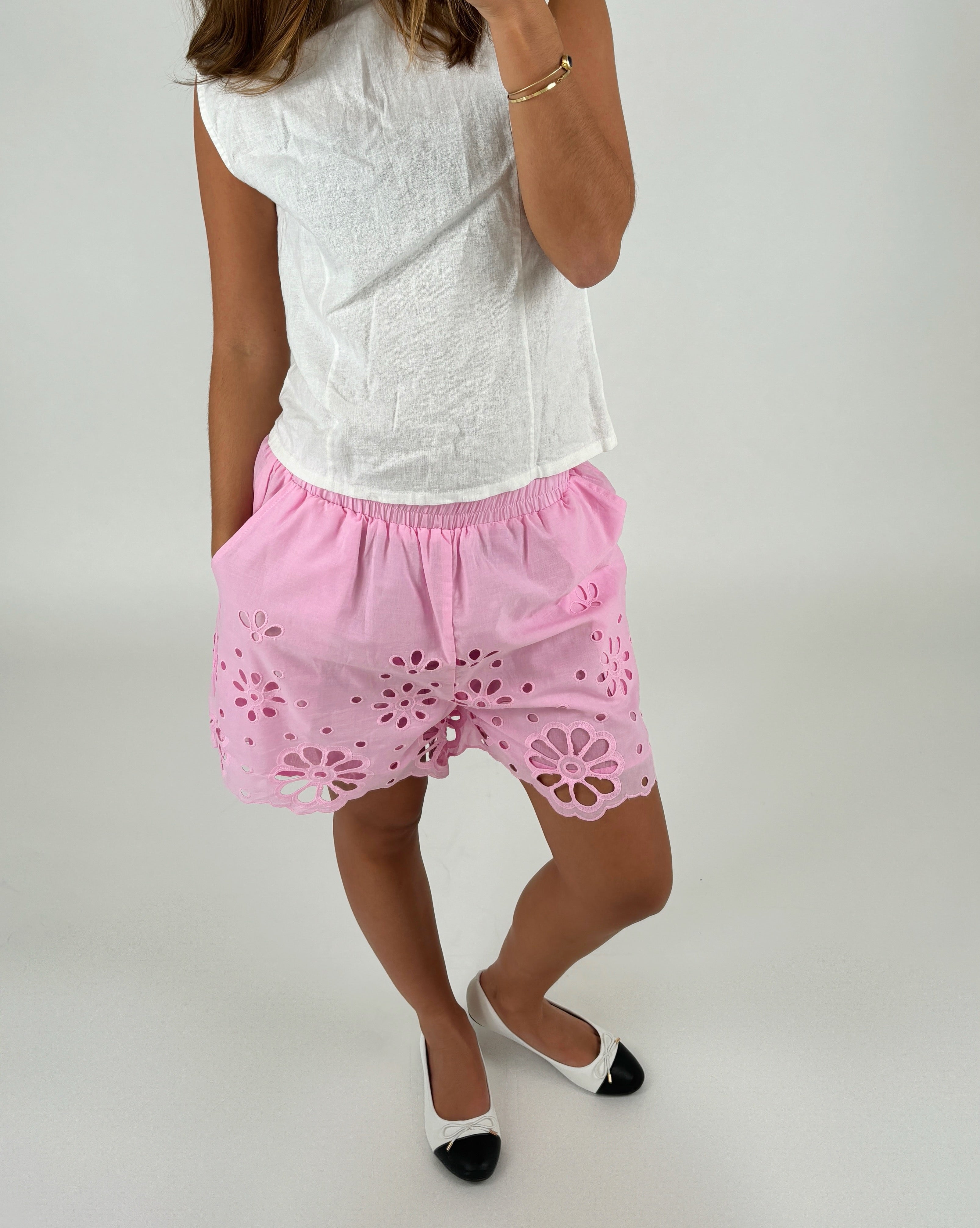 En person iført en hvid ærmeløs top og lyserøde shorts med blomstermønstre står i et neutralt rum. Personen bærer hvide sko med sorte snuder og et armbånd.