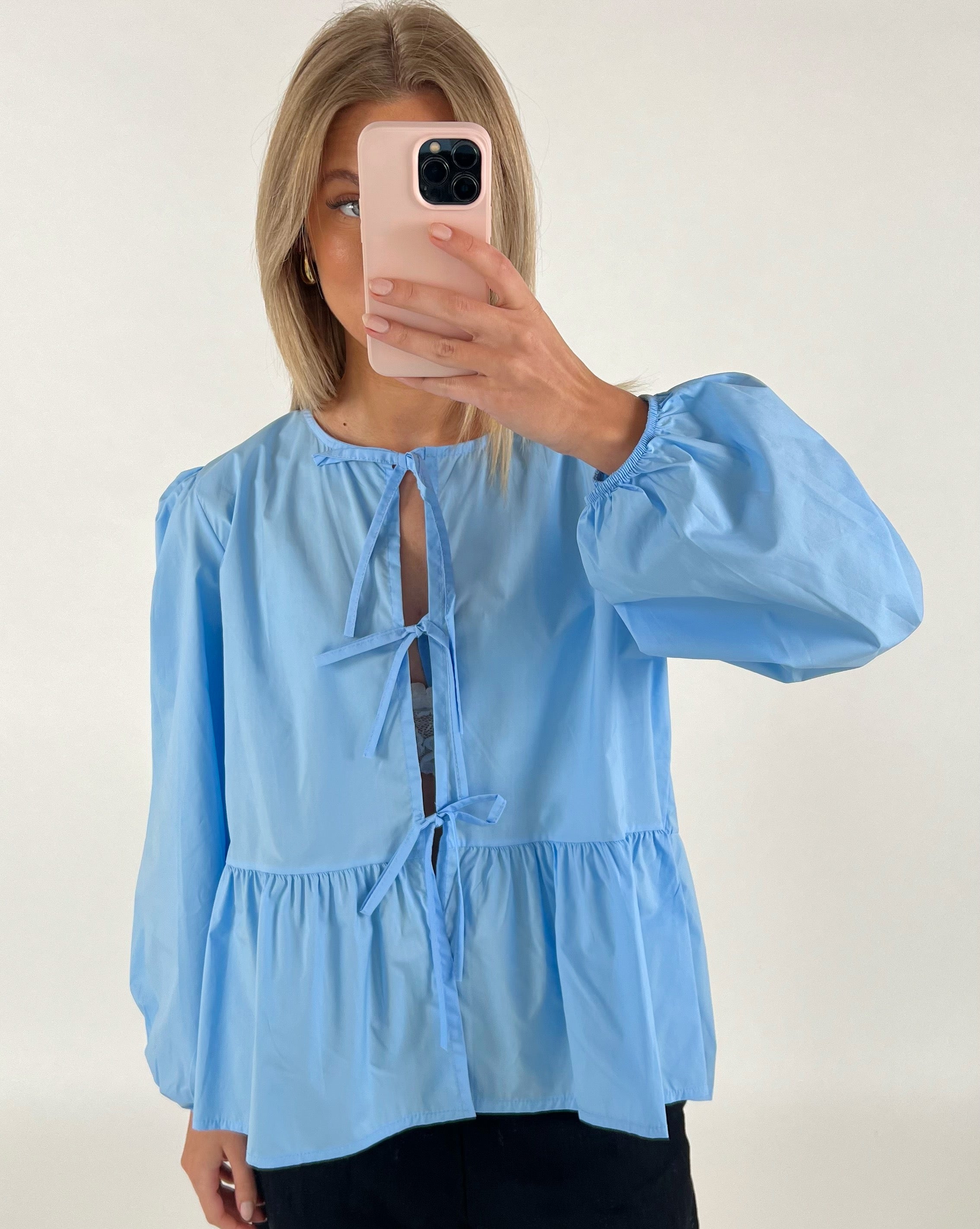 Person tager et spejlfoto med en smartphone iført en lyseblå bluse med bindebånd. Baggrunden er en minimalistisk, lys væg.