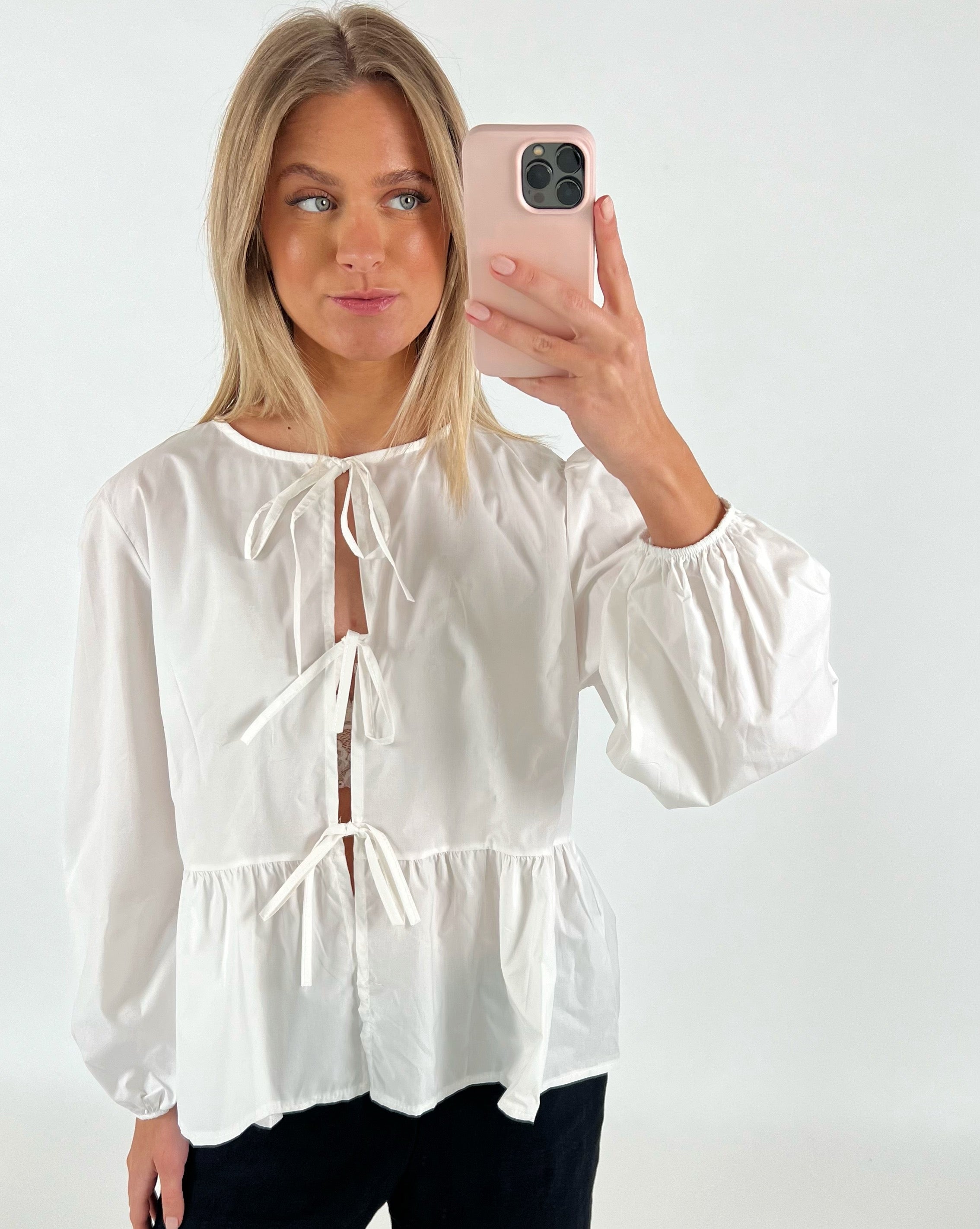 En kvinde iført en hvid bluse med sløjfer tager en selfie med sin smartphone. Hun står foran en ensfarvet, lys baggrund.