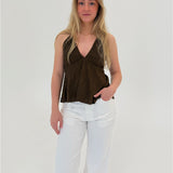 En person står stille iført en brun top og hvide bukser. Personen hviler en hånd i bukselommen i et lyst, minimalistisk rum.