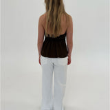 En person står med ryggen til, iført en stropløs brun top og hvide bukser, i et lyst studie uden synlige møbler eller dekorationer.