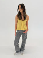 En kvinde står afslappet iført en gul top og grå bukser, med hånden i lommen. Hun befinder sig i et lyst, minimalistisk rum.