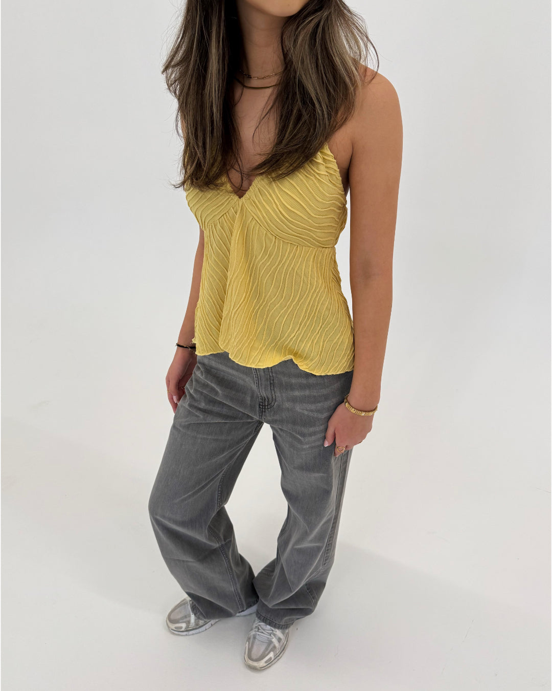 En person står iført en gul top og grå bukser, med hænderne afslappet ved siden, i et lyst, neutralt rum.