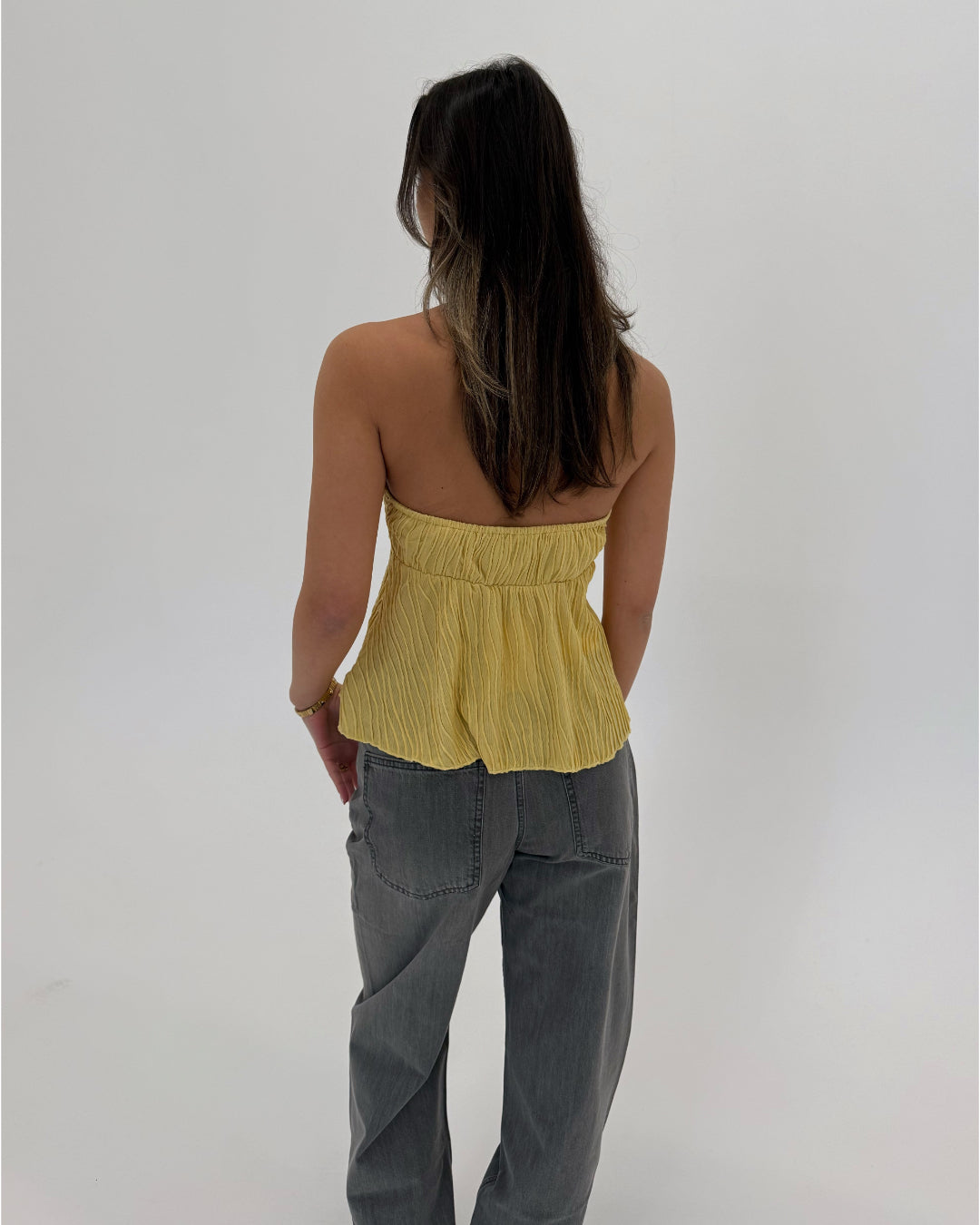 En person med langt mørkt hår står med ryggen til iført en gul stroptop og grå jeans. Baggrunden er helt hvid uden detaljer.