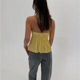 En person med langt mørkt hår står med ryggen til iført en gul stroptop og grå jeans. Baggrunden er helt hvid uden detaljer.