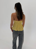 En person med langt mørkt hår står med ryggen til iført en gul stroptop og grå jeans. Baggrunden er helt hvid uden detaljer.