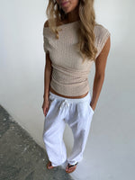 En person står iført en beige off-shoulder-top og hvide bukser med elastik i taljen. De står i et indendørs miljø med en grå betongulv og hvid væg.