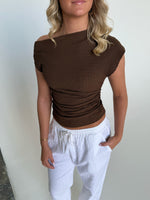 En person iført en brun, skulderfri top og hvide bukser står afslappet mod en hvid væg med en hånd i lommen.