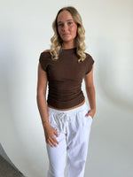 En person står afslappet iført en brun top og hvide bukser mod en enkel, hvid baggrund. Personen kigger direkte mod kameraet med hænderne i bukselommerne.