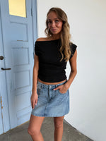 Kvinde står afslappet iført en sort top og denimnederdel. Hun har hånden i lommen. Baggrunden er en lys væg ved siden af en blå dør.