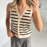 En person iført en ærmeløs, stribet vest med gyldne knapper tager et spejlbillede i et lyst, minimalistisk rum, iført grå bukser og et armbånd.