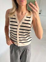 En person iført en ærmeløs, stribet vest med gyldne knapper tager et spejlbillede i et lyst, minimalistisk rum, iført grå bukser og et armbånd.