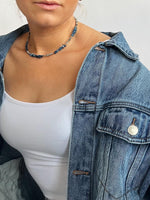 En person bærer en denimjakke over en hvid tanktop og en halskæde med blå perler; personen har lys hud og står mod en lys baggrund.