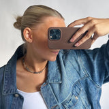 En person iført en denimjakke tager et billede med en mobiltelefon. Personen har opsat hår og bærer en hvid top og et perlekæde, set mod en lys baggrund.