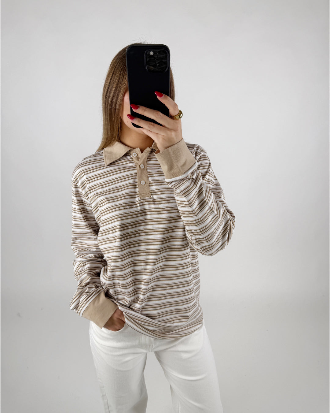 En person iført en stribet skjorte tager et selfie i et neutralt rum. Personen har hånden i lommen på et par hvide bukser.