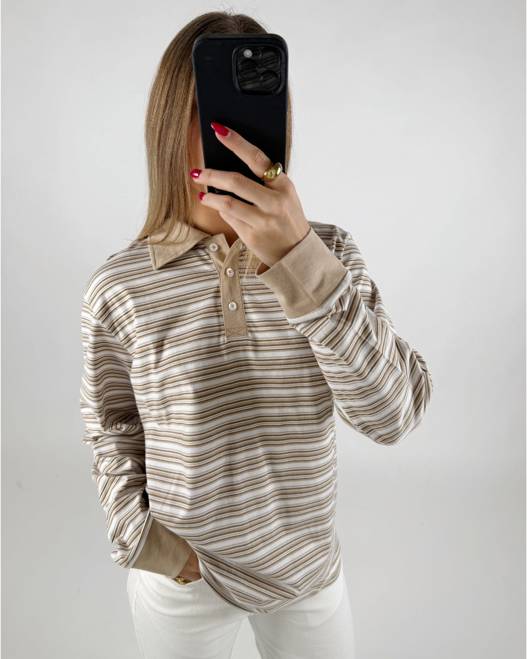 En person iført en stribet trøje tager et spejlselfie med en sort telefon. Personen har rødmalede negle og en guldring, i et neutralt belyst rum.
