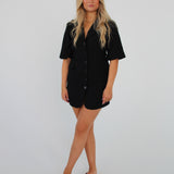En person står med armene afslappet, iført en sort kortærmet kjole og sandaler. Baggrunden er en ensfarvet, lys væg, hvilket giver et minimalistisk look.