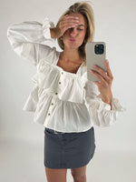 Person tager en selfie iført en hvid, detaljeret bluse og mørkt nederdel i et lyst rum. Personen har hånden over øjnene og laver et kysseansigt, mens mobilen fanger billedet.