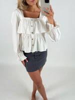 Kvinde tager et spejlbillede iført en hvid, knaplukket bluse med lange ærmer og en mørk nederdel. Hun står i et simpel hvidt rum, barfodet, med smartphonen i hånden.