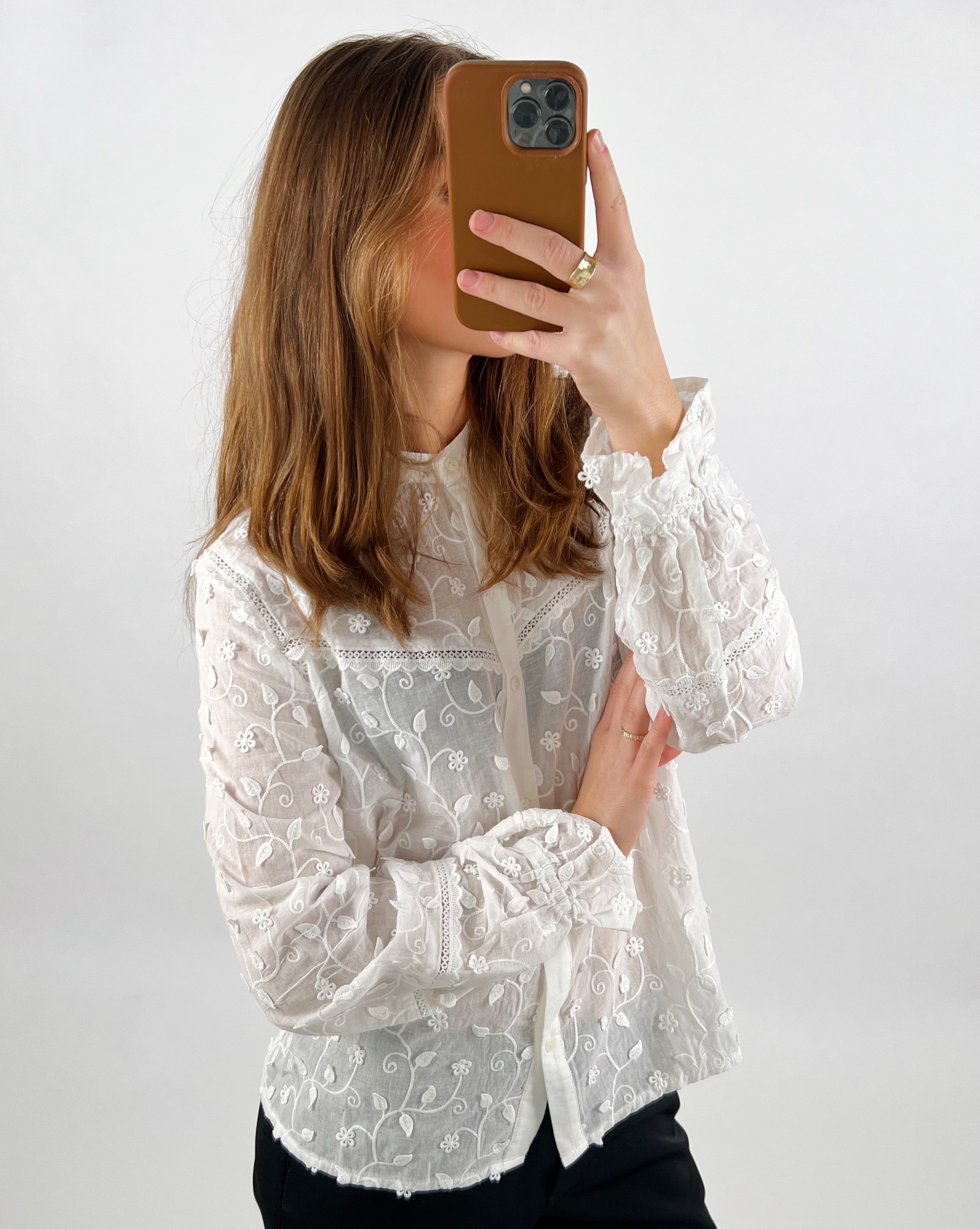 Person tager selfie med mobiltelefon iført hvid, broderet bluse. Baggrunden er enkel og lys, hvilket fremhæver det delikate mønster på blusen.