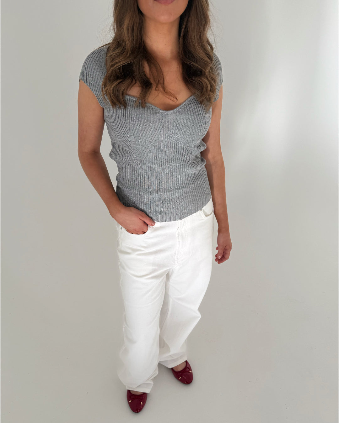 Kvinde iført grå top og hvide bukser står i et lyst rum, med én hånd i bukselommen. Hun har langt, bølget hår og bærer røde sko.