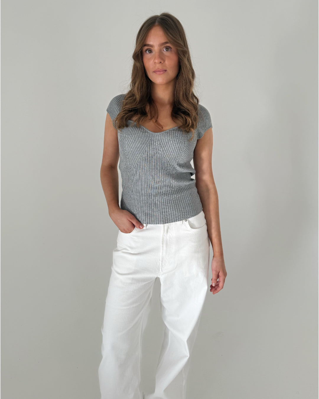 En kvinde står afslappet iført en grå strikket top og hvide bukser, med en neutral baggrund. Hendes ene hånd er i lommen.