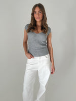 En kvinde står afslappet iført en grå strikket top og hvide bukser, med en neutral baggrund. Hendes ene hånd er i lommen.