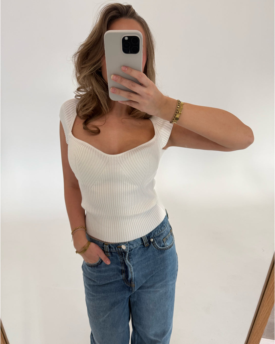 En person tager et spejlbillede med en smartphone, iført en hvid ærmeløs top og blå jeans, i et lyst rum.