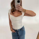 En person tager et spejlbillede med en smartphone, iført en hvid ærmeløs top og blå jeans, i et lyst rum.