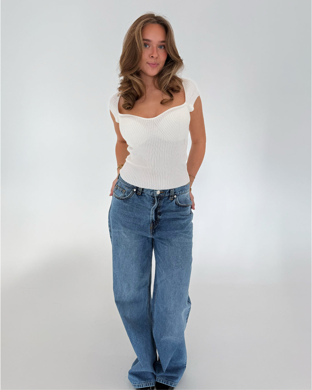Kvinde står afslappet i hvid top og blå jeans i et lyst rum. Hun har hånden i lommen og blikket rettet mod kameraet.
