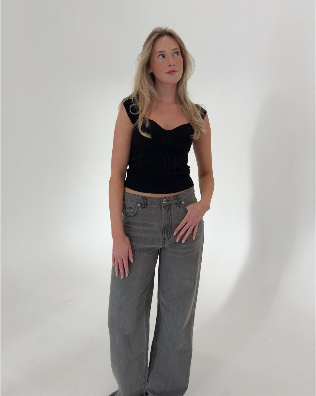 En kvinde i sort top og grå bukser står afslappet med hænderne i lommen, i et lyst, minimalistisk rum. Hun ser opad, med let bølget hår.