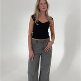 En kvinde i sort top og grå bukser står afslappet med hænderne i lommen, i et lyst, minimalistisk rum. Hun ser opad, med let bølget hår.