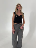 En kvinde i sort top og grå bukser står afslappet med hænderne i lommen, i et lyst, minimalistisk rum. Hun ser opad, med let bølget hår.