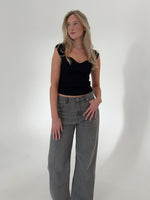 En kvinde står, iført en sort top og grå bukser, ser til siden i et minimalistisk, hvidt rum.