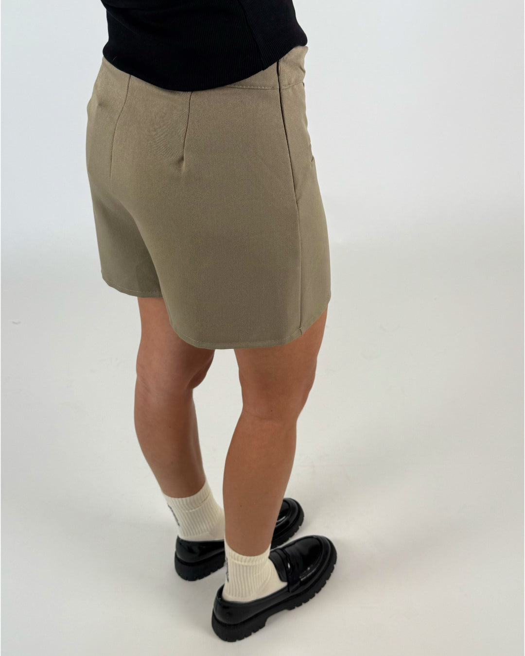 En person iført beige shorts står oprejst, iført sorte sko og hvide strømper, på en lys baggrund. Personen bærer også en sort trøje.