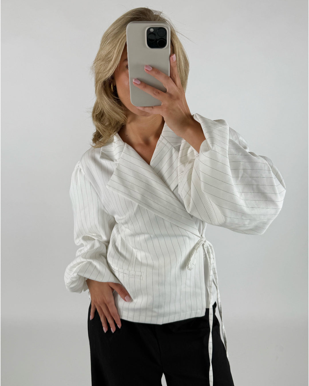 En person tager en selfie med smartphone og dækker ansigtet, iført en hvid, stribet bluse med lange ærmer. Baggrunden er ensfarvet lys.