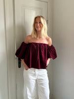 En person står smilende iført en mørkerød, skulderløs top og hvide bukser, med hænderne i lommerne, foran en hvid dør i et lyst rum.