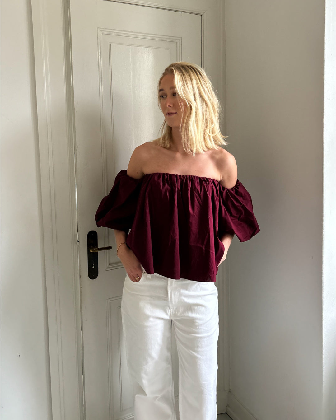 En person står i en gang iført en burgundy off-shoulder bluse og hvide bukser. Baggrunden viser en hvid dør og vægge.